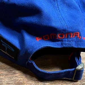 Puede incluir: Una gorra de b&eacute;isbol azul con la palabra "POMONA, CA" bordada en rojo en la parte trasera. La gorra tiene una visera curva y una correa ajustable. Una etiqueta negra con la palabra "POWERANG" est&aacute; en el lateral.