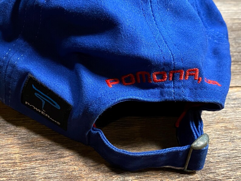 Puede incluir: Una gorra de b&eacute;isbol azul con la palabra "POMONA, CA" bordada en rojo en la parte trasera. La gorra tiene una visera curva y una correa ajustable. Una etiqueta negra con la palabra "POWERANG" est&aacute; en el lateral.