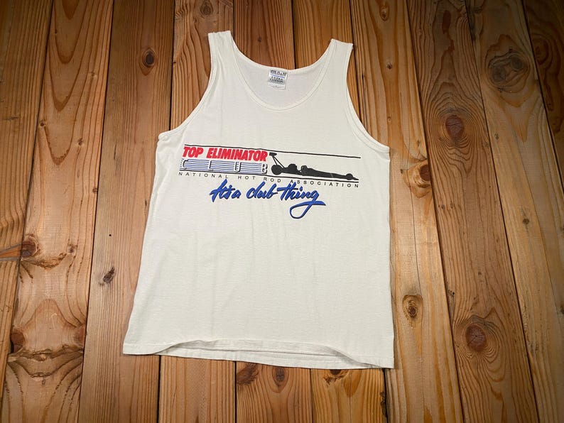 May include: White tank top with the text "TOP ELIMINATOR CLUB National Hot Rod Association It's a club thing" in red, blue, and black. The tank top is made of a soft, lightweight material.