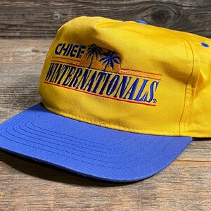 May include: A yellow baseball cap with a blue brim. The cap features the text "CHIEF WINTERNATIONALS" in blue and red, with palm tree graphics. The hat is made of a durable fabric.