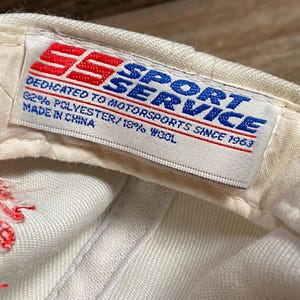 Op de afbeelding: Close-up van de binnenkant van een witte baseballcap met een label. Het label bevat de tekst "SPORT SERVICE" in blauw, met "SS" in rood. Er staat "DEDICATED TO MOTORSPORTS SINCE 1963" en "82% POLYESTER/18% WOOL. Made in China."