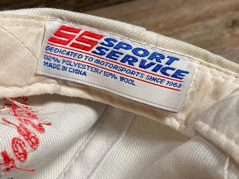 Op de afbeelding: Close-up van de binnenkant van een witte baseballcap met een label. Het label bevat de tekst "SPORT SERVICE" in blauw, met "SS" in rood. Er staat "DEDICATED TO MOTORSPORTS SINCE 1963" en "82% POLYESTER/18% WOOL. Made in China."