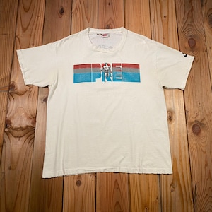 以下が含まれることがあります： クリーム色のTシャツ。グラフィックデザインは、赤、青、白のストライプが入った長方形のブロックに「PRIDE」の文字が描かれています。袖には小さなナイキのロゴが見えます。木製の表面に平らに置かれたシャツ。