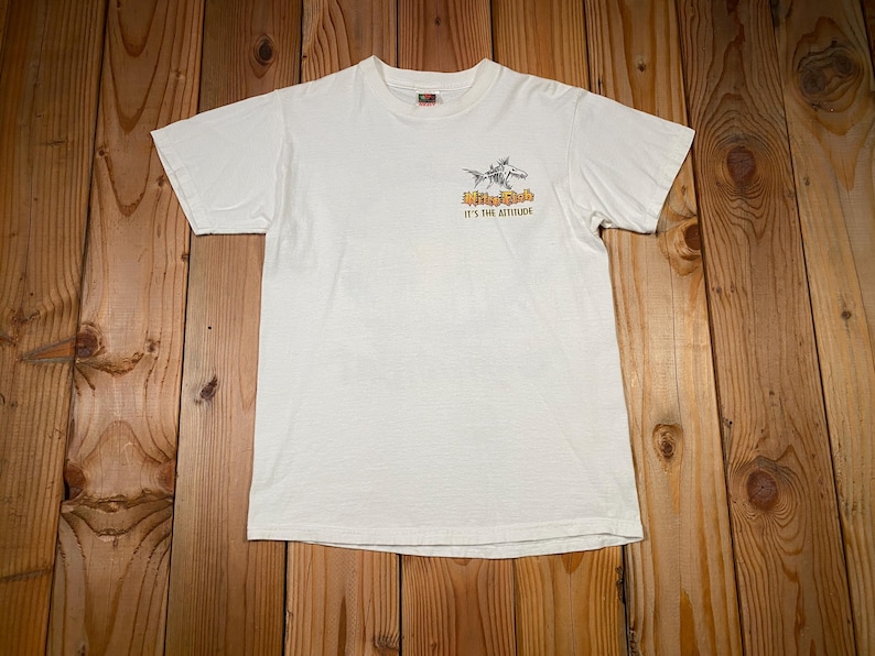 Puede incluir: Camiseta blanquecina con un peque&ntilde;o gr&aacute;fico en la parte delantera. El gr&aacute;fico presenta un pez estilizado y el texto "Mike Fish It's the Attitude" en naranja y negro. La camiseta es de un material suave.