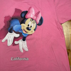 May include: A bright pink t-shirt featuring a graphic of Minnie Mouse emerging from a torn fabric effect. Minnie wears a pink bow and blue clothing. The word California is printed in white text below the graphic on the front of the shirt.