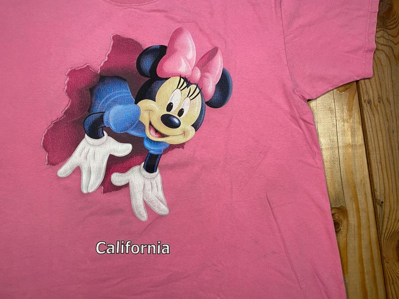 May include: A bright pink t-shirt featuring a graphic of Minnie Mouse emerging from a torn fabric effect. Minnie wears a pink bow and blue clothing. The word California is printed in white text below the graphic on the front of the shirt.
