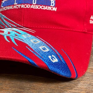 Op de afbeelding: Een rode baseballpet met geborduurde blauwe en zilveren vlamachtige graphics en de tekst 2003. De pet toont het logo van de National Hot Rod Association en aanvullende tekst, waaronder Automobile Southern California NHRA op het zijpaneel.