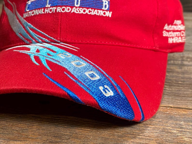 Op de afbeelding: Een rode baseballpet met geborduurde blauwe en zilveren vlamachtige graphics en de tekst 2003. De pet toont het logo van de National Hot Rod Association en aanvullende tekst, waaronder Automobile Southern California NHRA op het zijpaneel.