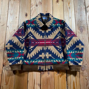 May include: A zip-up jacket featuring a geometric pattern in navy blue, tan, red, and teal. The garment has a fold-down collar, long sleeves with ribbed cuffs, and a ribbed hem. A brown interior lining is visible at the collar and a label is present.