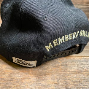 May include: Black baseball cap with a flat brim and adjustable snapback closure. The words "MEMBERS ONLY" are embroidered in a light green color on the back of the cap. A small white tag with the text "TORX ELIMINATOR" is on the side.