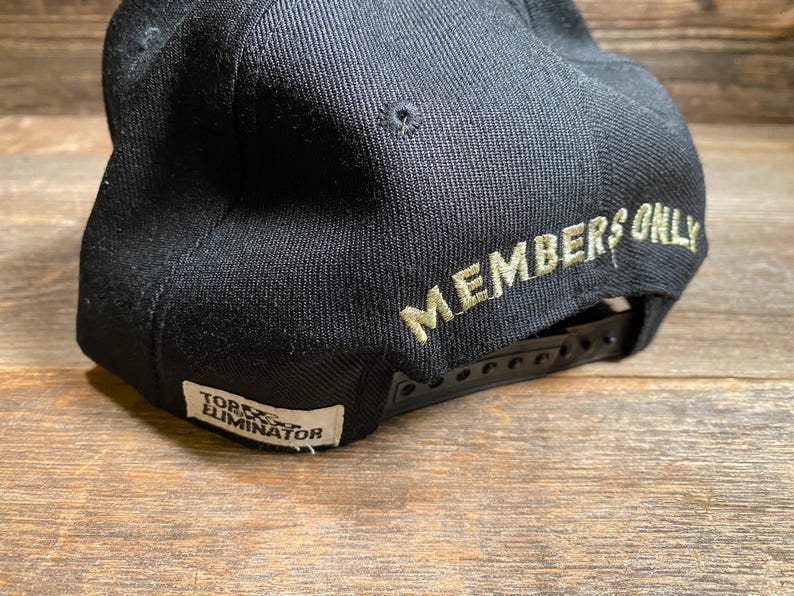 May include: Black baseball cap with a flat brim and adjustable snapback closure. The words "MEMBERS ONLY" are embroidered in a light green color on the back of the cap. A small white tag with the text "TORX ELIMINATOR" is on the side.