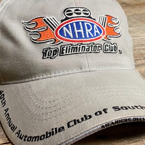 May include: A light grey baseball cap with the NHRA logo in the centre, featuring a red, white, and blue oval with the text "NHRA". The cap also has the text "Top Eliminator Club" and "40th Annual Automobile Club of Southern California".