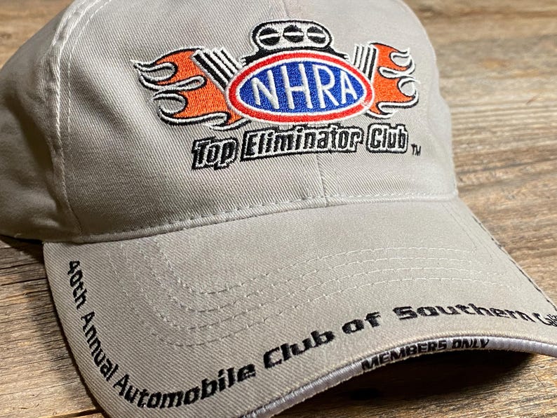 May include: A light grey baseball cap with the NHRA logo in the centre, featuring a red, white, and blue oval with the text "NHRA". The cap also has the text "Top Eliminator Club" and "40th Annual Automobile Club of Southern California".