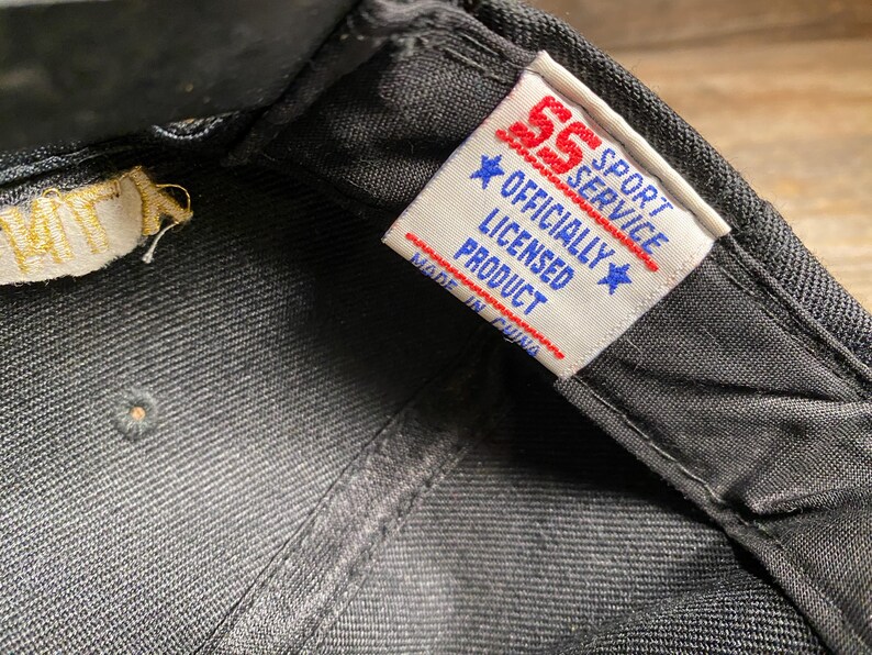 May include: Close-up of a black baseball cap. The interior features a tag with the text "SS Sport Service Officially Licensed Product Made in China". The tag has red and blue accents.