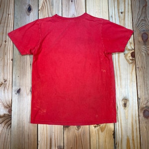 Op de afbeelding: Een effen rood T-shirt met een ronde hals en korte mouwen. Het shirt heeft een licht vervaagde uitstraling. Het T-shirt wordt gepresenteerd op een houten oppervlak.