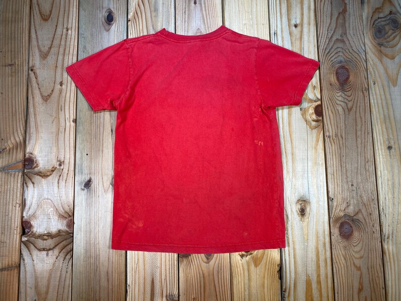 Op de afbeelding: Een effen rood T-shirt met een ronde hals en korte mouwen. Het shirt heeft een licht vervaagde uitstraling. Het T-shirt wordt gepresenteerd op een houten oppervlak.