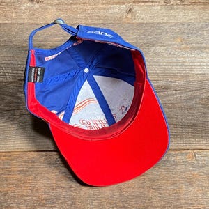 Puede incluir: Gorra de b&eacute;isbol azul con visera roja e interior blanco. La gorra tiene la palabra "zone" en la parte delantera y una etiqueta que dice "Manufactured in Insert Japan". La gorra est&aacute; sobre una superficie de madera.