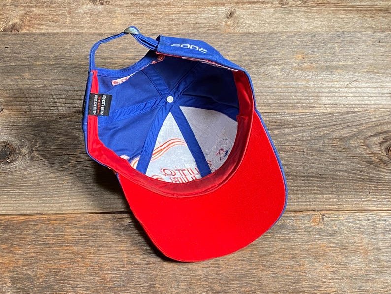 Puede incluir: Gorra de b&eacute;isbol azul con visera roja e interior blanco. La gorra tiene la palabra "zone" en la parte delantera y una etiqueta que dice "Manufactured in Insert Japan". La gorra est&aacute; sobre una superficie de madera.