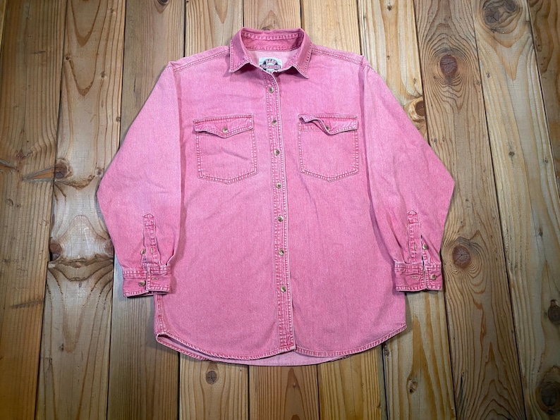 Puede incluir: Una camisa de manga larga con botones en color rosa deste&ntilde;ido. La camisa presenta un cuello cl&aacute;sico, dos bolsillos en el pecho con solapas abotonadas y botones de metal en la parte delantera y en los pu&ntilde;os. Una peque&ntilde;a etiqueta blanca est&aacute; en el cuello.