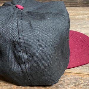 May include: A black baseball cap with a maroon brim and button. The cap is made of a textured fabric and has a classic, structured design. The brim is curved and the button is maroon.