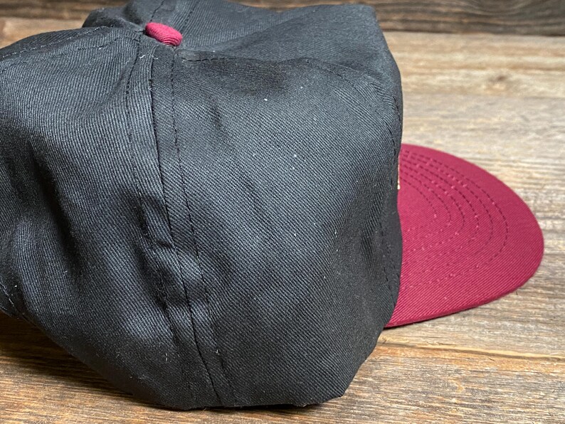 May include: A black baseball cap with a maroon brim and button. The cap is made of a textured fabric and has a classic, structured design. The brim is curved and the button is maroon.