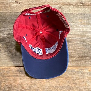 May include: A red baseball cap with a dark blue brim, viewed from the inside. The interior features red fabric panels, a white button at the crown, and white interior seam tape with blue text that reads MINATOR 2010.