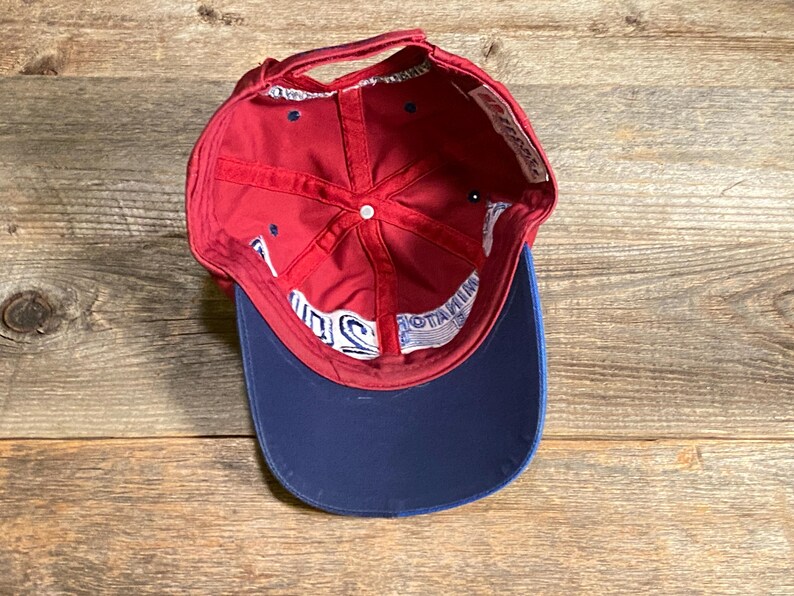 May include: A red baseball cap with a dark blue brim, viewed from the inside. The interior features red fabric panels, a white button at the crown, and white interior seam tape with blue text that reads MINATOR 2010.