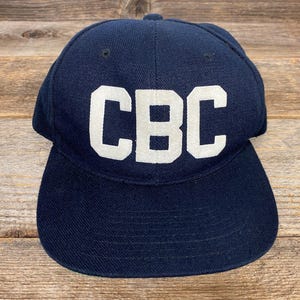 Könnte beinhalten: Eine marineblaue Baseballkappe mit den weißen Buchstaben "CBC" auf der Vorderseite. Die Kappe hat einen gebogenen Schirm und einen Knopf oben. Der Hut ist aus einem strukturierten Stoff gefertigt und liegt auf einer Holzoberfläche.