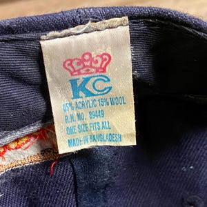 May include: A navy blue baseball cap interior featuring a white fabric tag with a red crown icon and blue text reading KC, 85% ACRYLIC 15% WOOL, R.N. NO. 89449, ONE SIZE FITS ALL, and MADE IN BANGLADESH, with visible red and gold decorative stitching.