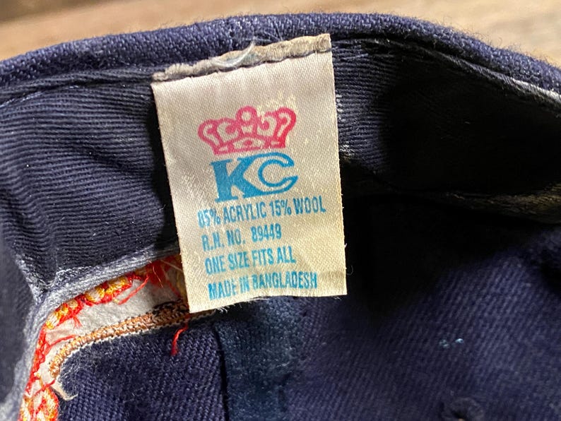 May include: A navy blue baseball cap interior featuring a white fabric tag with a red crown icon and blue text reading KC, 85% ACRYLIC 15% WOOL, R.N. NO. 89449, ONE SIZE FITS ALL, and MADE IN BANGLADESH, with visible red and gold decorative stitching.