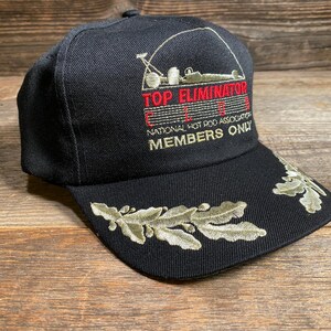 May include: Black baseball cap with embroidered text "TOP ELIMINATOR" and "NATIONAL HOT ROD ASSOCIATION MEMBERS ONLY." Features a leaf design on the brim. A classic hat for motorsports enthusiasts.