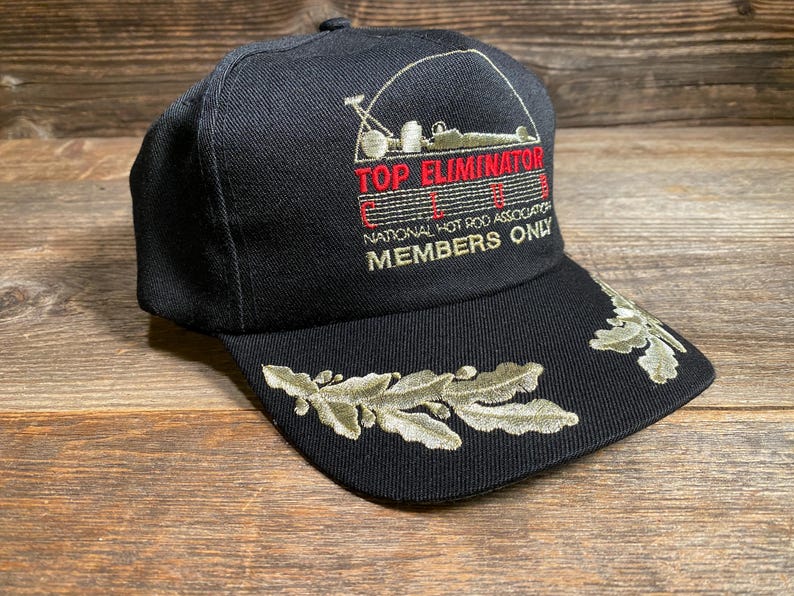 May include: Black baseball cap with embroidered text "TOP ELIMINATOR" and "NATIONAL HOT ROD ASSOCIATION MEMBERS ONLY." Features a leaf design on the brim. A classic hat for motorsports enthusiasts.
