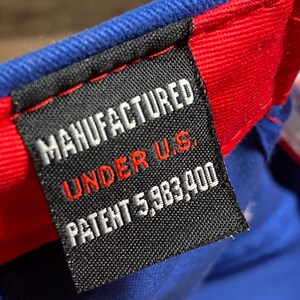 Puede incluir: Primer plano de una etiqueta en una gorra de b&eacute;isbol azul y roja. La etiqueta dice "MANUFACTURED UNDER U.S. PATENT 5,983,400". La etiqueta es negra con texto blanco y rojo.
