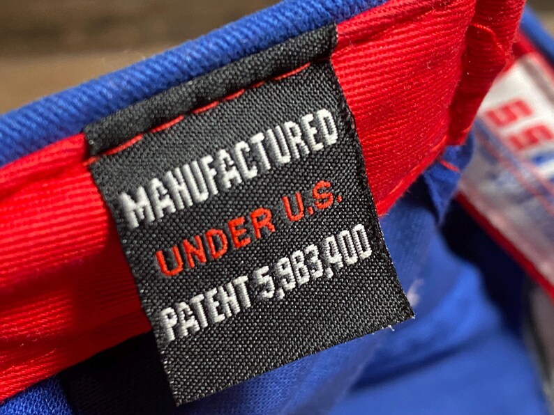Puede incluir: Primer plano de una etiqueta en una gorra de b&eacute;isbol azul y roja. La etiqueta dice "MANUFACTURED UNDER U.S. PATENT 5,983,400". La etiqueta es negra con texto blanco y rojo.