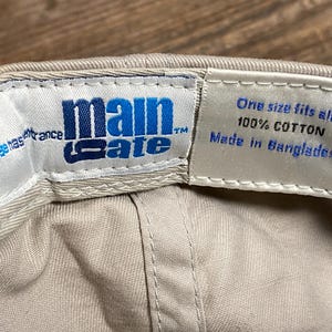 May include: A beige baseball cap with the blue text "main gate". The interior label states "One size fits all 100% COTTON Made in Bangladesh". The cap is made of cotton.