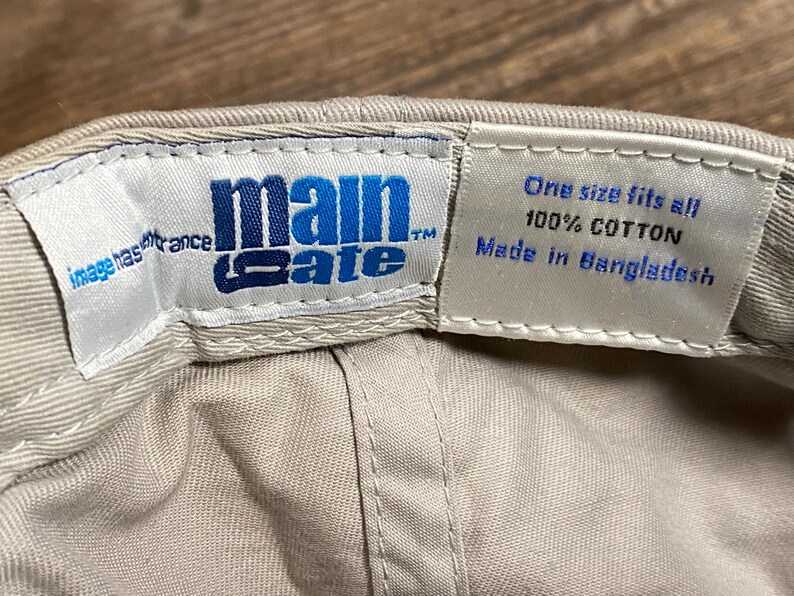 May include: A beige baseball cap with the blue text "main gate". The interior label states "One size fits all 100% COTTON Made in Bangladesh". The cap is made of cotton.