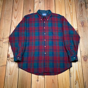 Puede incluir: Una camisa de cuadros de manga larga en tonos rojos, verde azulado y marrón. La camisa con botones tiene cuello y un bolsillo en el pecho izquierdo. La camisa está hecha de un material suave y cómodo.