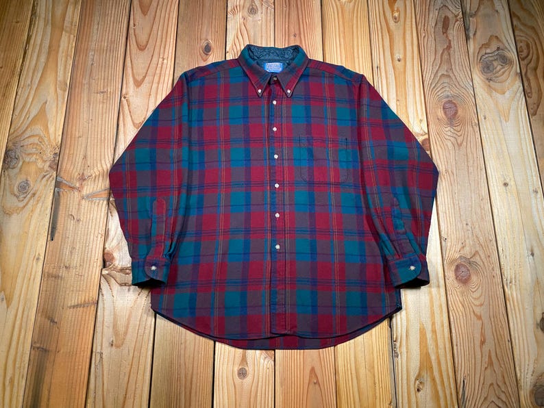 May include: A long-sleeved plaid shirt in shades of red, teal, and brown. The button-down shirt has a collared neckline and a pocket on the left chest. The shirt is made of a soft, comfortable material.