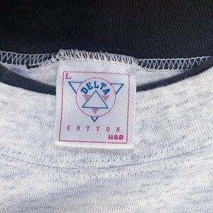 May include: A gray t-shirt with a black collar and a white tag that says "DELTA" in a triangle with the words "COTTON USA" below it. The tag has a size "L" printed on it.