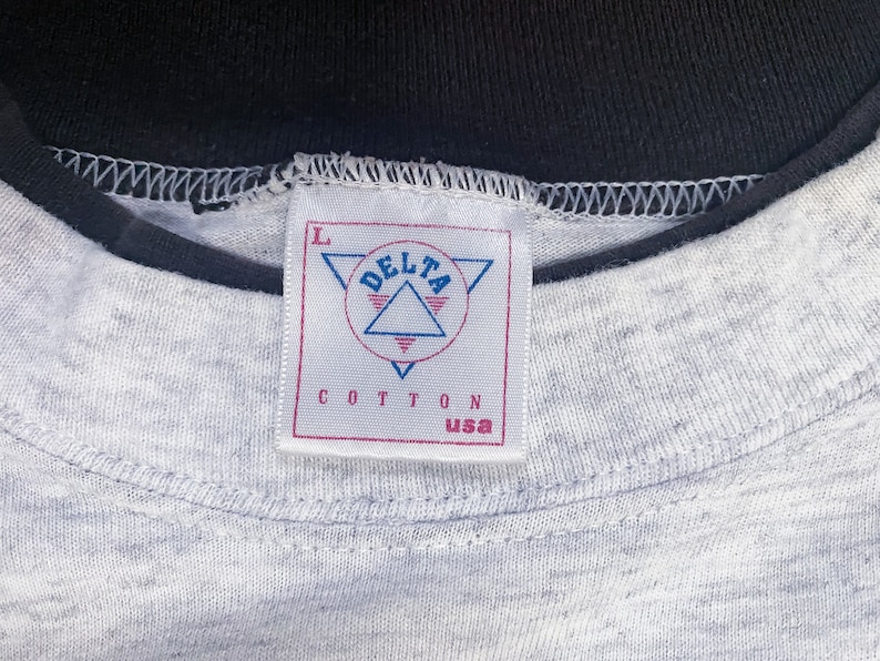 May include: A gray t-shirt with a black collar and a white tag that says "DELTA" in a triangle with the words "COTTON USA" below it. The tag has a size "L" printed on it.