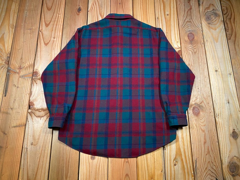 May include: A long-sleeved plaid shirt in a classic pattern. The shirt features a button-down collar and cuffs. The plaid design incorporates shades of burgundy, teal, and navy blue. The shirt is made of a soft, possibly flannel, material.