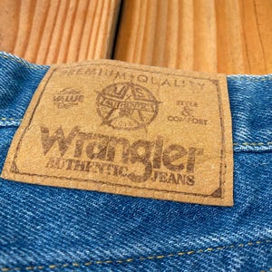 May include: Close-up of blue denim fabric with a brown leather patch. The patch reads "Wrangler AUTHENTIC JEANS" with additional text and a star emblem. The jeans are on a wooden surface.