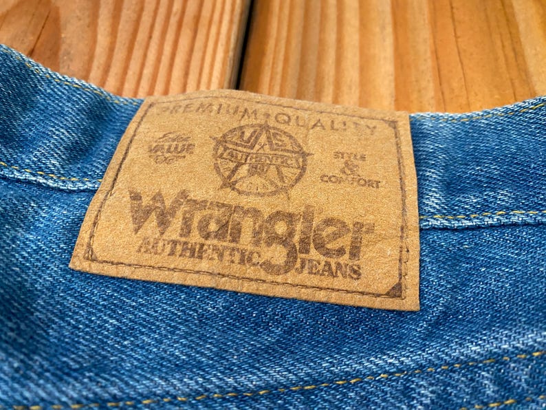 May include: Close-up of blue denim fabric with a brown leather patch. The patch reads "Wrangler AUTHENTIC JEANS" with additional text and a star emblem. The jeans are on a wooden surface.
