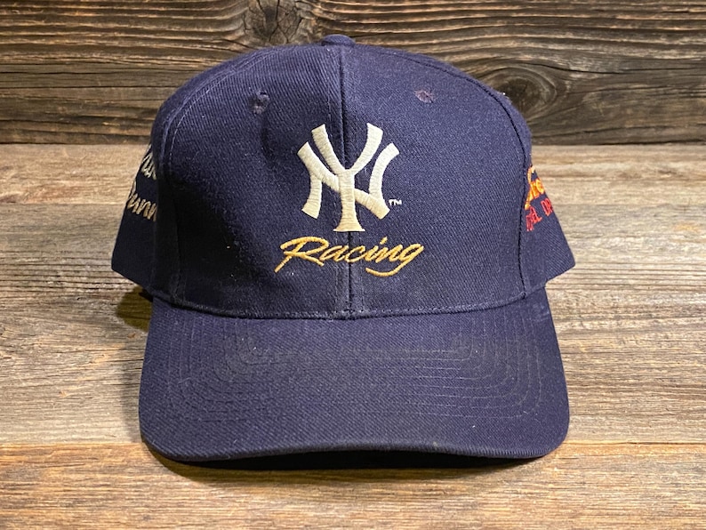 May include: A navy blue baseball cap featuring an embroidered white New York Yankees logo on the front with the word Racing in gold script underneath. The cap includes additional embroidered text details on the side panels in gold and white thread.