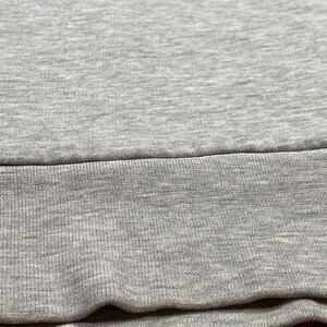 Puede incluir: Primer plano de una tela gris, probablemente una sudadera o prenda similar. La imagen muestra la textura del material, con un dobladillo acanalado en la parte inferior. La tela parece suave y c&oacute;moda, adecuada para ropa informal.