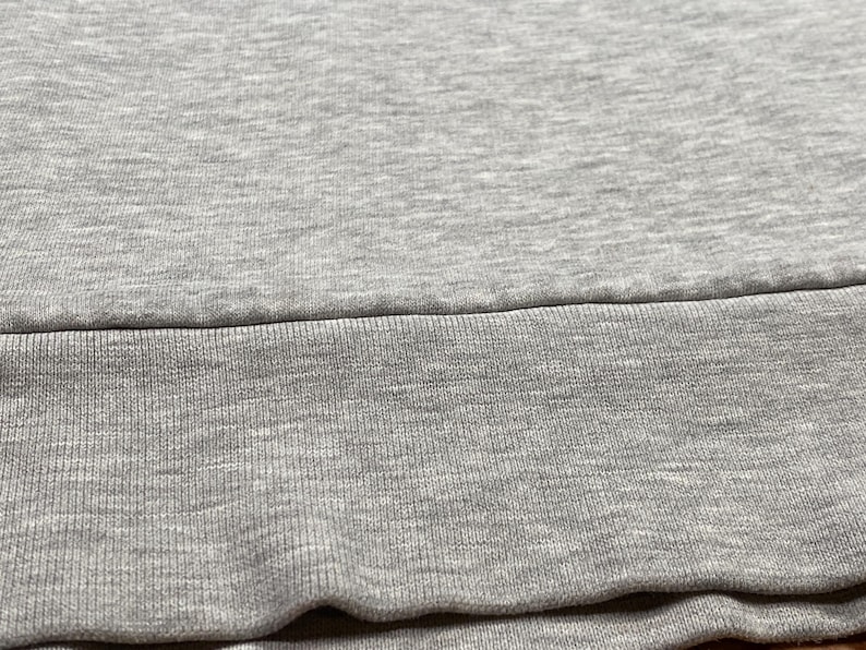 Puede incluir: Primer plano de una tela gris, probablemente una sudadera o prenda similar. La imagen muestra la textura del material, con un dobladillo acanalado en la parte inferior. La tela parece suave y c&oacute;moda, adecuada para ropa informal.