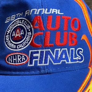 Puede incluir: Gorra de b&eacute;isbol azul con el texto "38th Annual Auto Club Finals" en rojo y blanco. La gorra presenta el logotipo de AAA Southern California Automobile Club y el logotipo de NHRA. Llamas en amarillo y naranja adornan el lateral.
