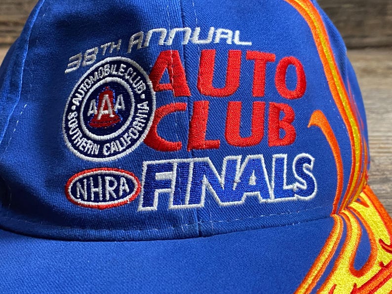 Puede incluir: Gorra de b&eacute;isbol azul con el texto "38th Annual Auto Club Finals" en rojo y blanco. La gorra presenta el logotipo de AAA Southern California Automobile Club y el logotipo de NHRA. Llamas en amarillo y naranja adornan el lateral.