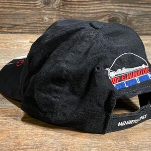 May include: Black baseball cap with the text "TOP ELIMINATOR CLUB" in red, white, and blue. The cap has a curved brim and an adjustable strap with the words "MEMBERS ONLY".