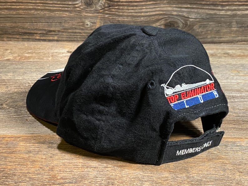 May include: Black baseball cap with the text "TOP ELIMINATOR CLUB" in red, white, and blue. The cap has a curved brim and an adjustable strap with the words "MEMBERS ONLY".